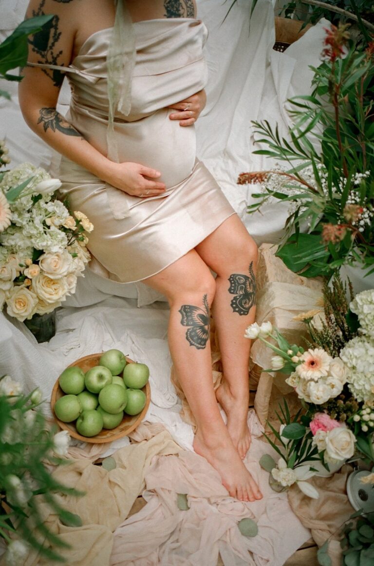 Hands resting on a pregnant belly beside a bowl of green apples and flowers