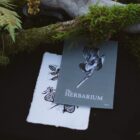 The Herbarium Vol. 1 and a hand-carved linocut plant card resting beneath a mossy branch