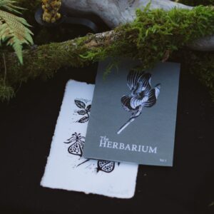The Herbarium Vol. 1 and a hand-carved linocut plant card resting beneath a mossy branch